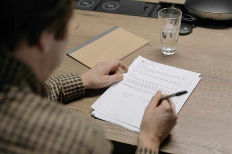 Un homme, vu de dos et en plongée, signe un contrat au format papier déposé sur une table en bois, sur laquelle se trouve un carnet et un verre d’eau